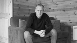 Black and white picture of a man sitting on a sofa leaning forward with a serious look on his face. His arms are resting on his knees and his hands are clasped together.