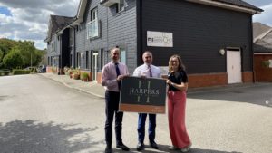 Two men and a woman smiling as they hold up a small advertising board that says Harpers. They are standing outside in front of a large wooden office building.