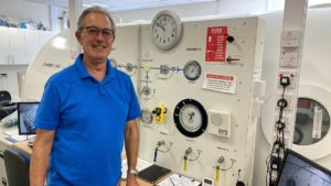 Smiling man standing in fron of a large white metal chamber. The panel on the chamber contains a series of dials and and levers.