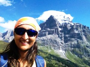 Smiling young lady wearing sunglasses and a bandana standing in front of high mountains.