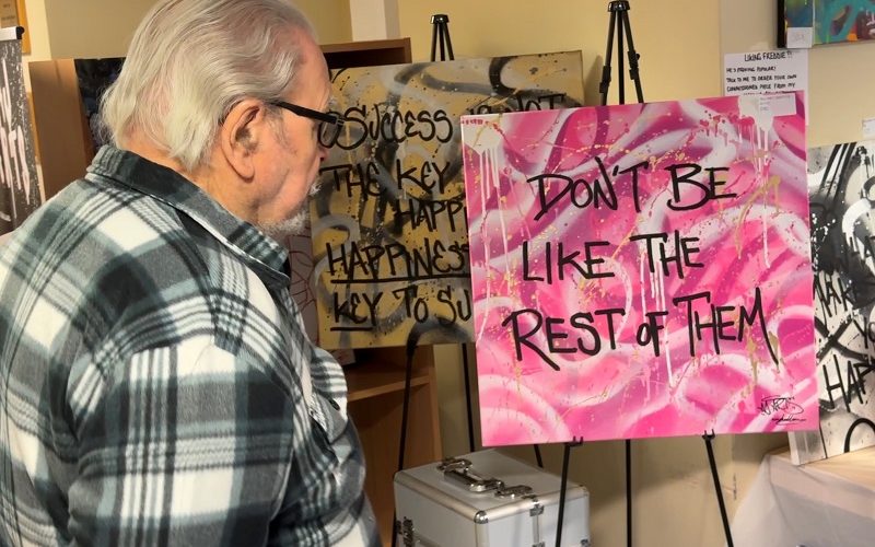 View of an art exhibition with a man looking intently at a piece of grafitti art which is bright pink with text saying don't be like the rest of them