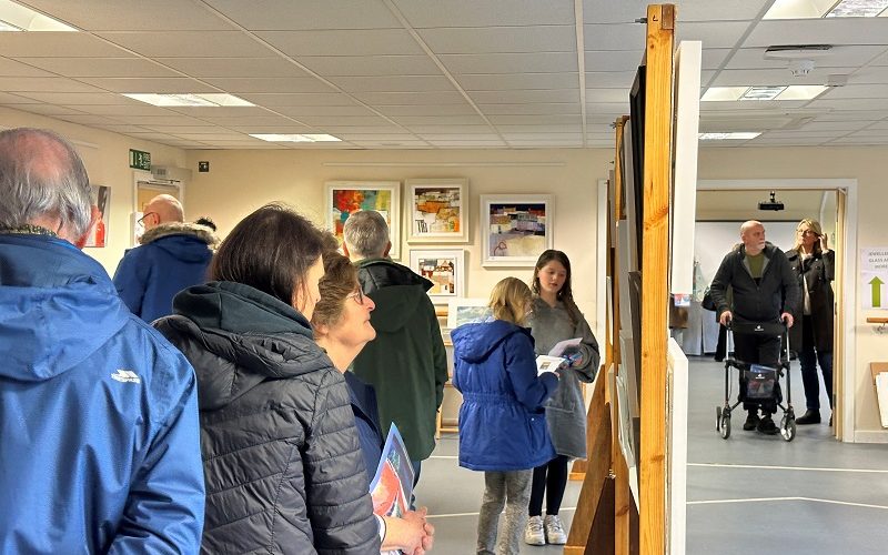 View of a busy art exhibition with paintings on the wall and on central frames.