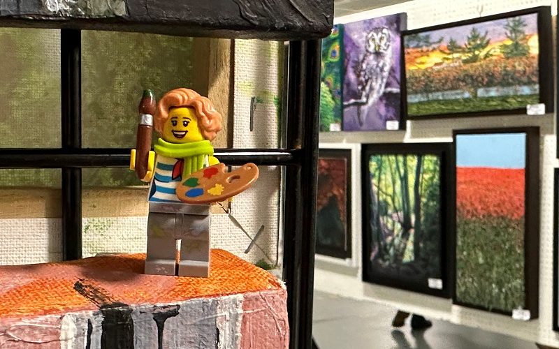View of an art exhibition with several paintings in the background. In the foreground is a Lego minifigure holding a paintbrush and palette