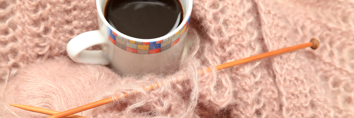 Mug of black coffee sitting among of a pile of wool and a woolly scarf with two knitting needles sticking out.