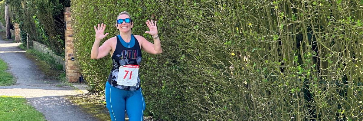 Woman taking part in a running race wearing sunglasses and a bib with number 71 on it. She has a big smile on her face and is waving her hands in the air.