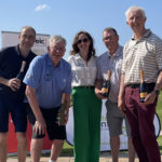 Four men standing two either side of a lady at a golf course. They are all smiling because their team has won. The woman in the middle has just presented them with their prizes - a bottle of Prosecco - for being the winning team.