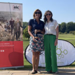 Two women standing outside at a golf course in front of a banner for EIC Insurance. Both our smiling. The person on the left is being presented with their prize - a bottle of Prosecco - by the person on the right.