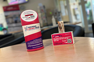 A bright pink collection tin for the Chilterns Neuro Centre sitting on a table in a large open reception area next to a small cardboard collection box, also in bright pink. In the collection box there is a £10 note sticking out.