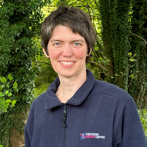 Headshot of smiling lady with short dark hair. She is standing in front of a forested area and is wearing a navy polo short with the logo for the Chilterns Neuro Centre.