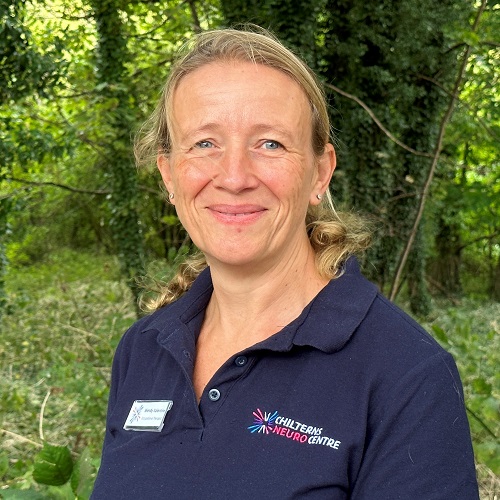 Headshot of smiling lady with fair hair. She is standing in front of a forested area and is wearing a navy polo short with the logo for the Chilterns Neuro Centre.