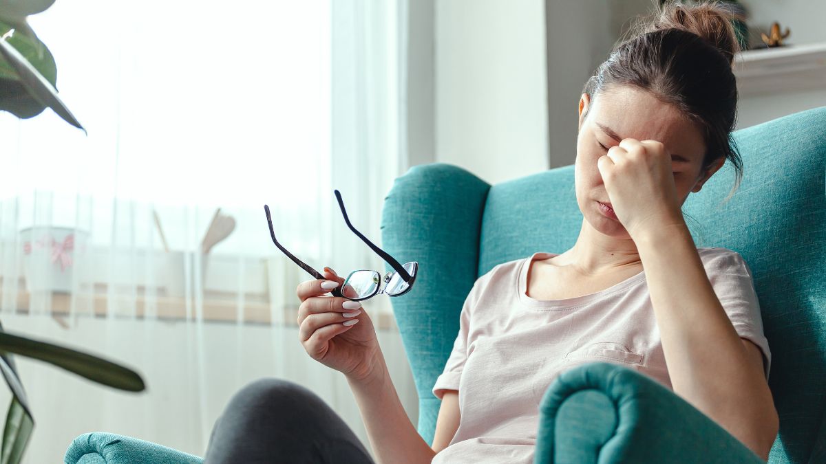 Woman sitting on a sofa holding her glasses in one hand and rubbing her eyes with the other. she is looking completely exhausted.