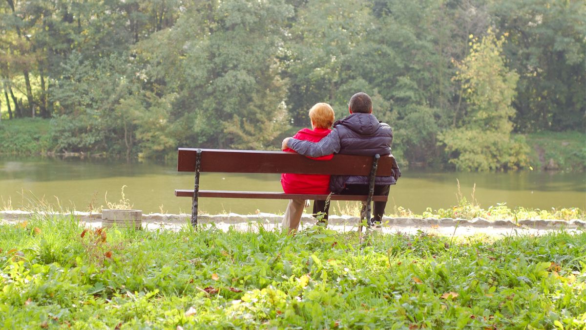 Couple sitting on a bench by the riverside, with their arms around each other.