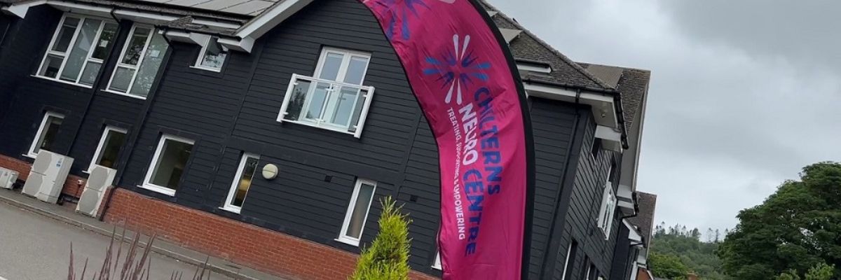 Corner of a large wooden building and in the foreground is a tall pink feather flag with the logo for the Chilterns Neuro Centre.