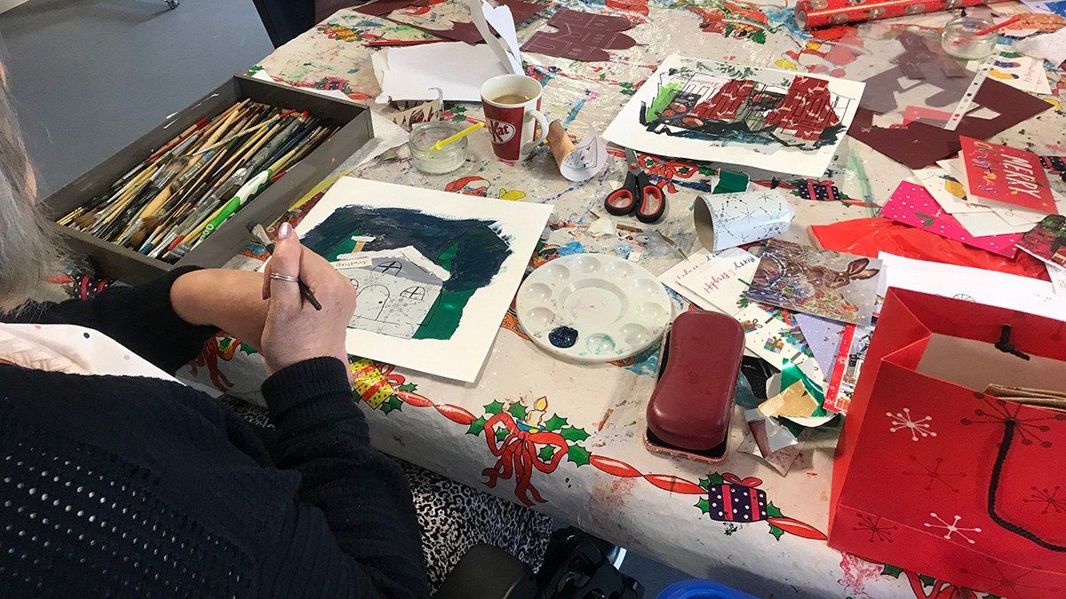 Over the shoulder shot of a lady painting a icture. The table she is sitting at is covered with paper and brushes.