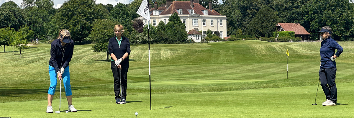 Lady on a golf course about to sink her putt while being watched by her her two friends. In the background is a large white-washed grand house.
